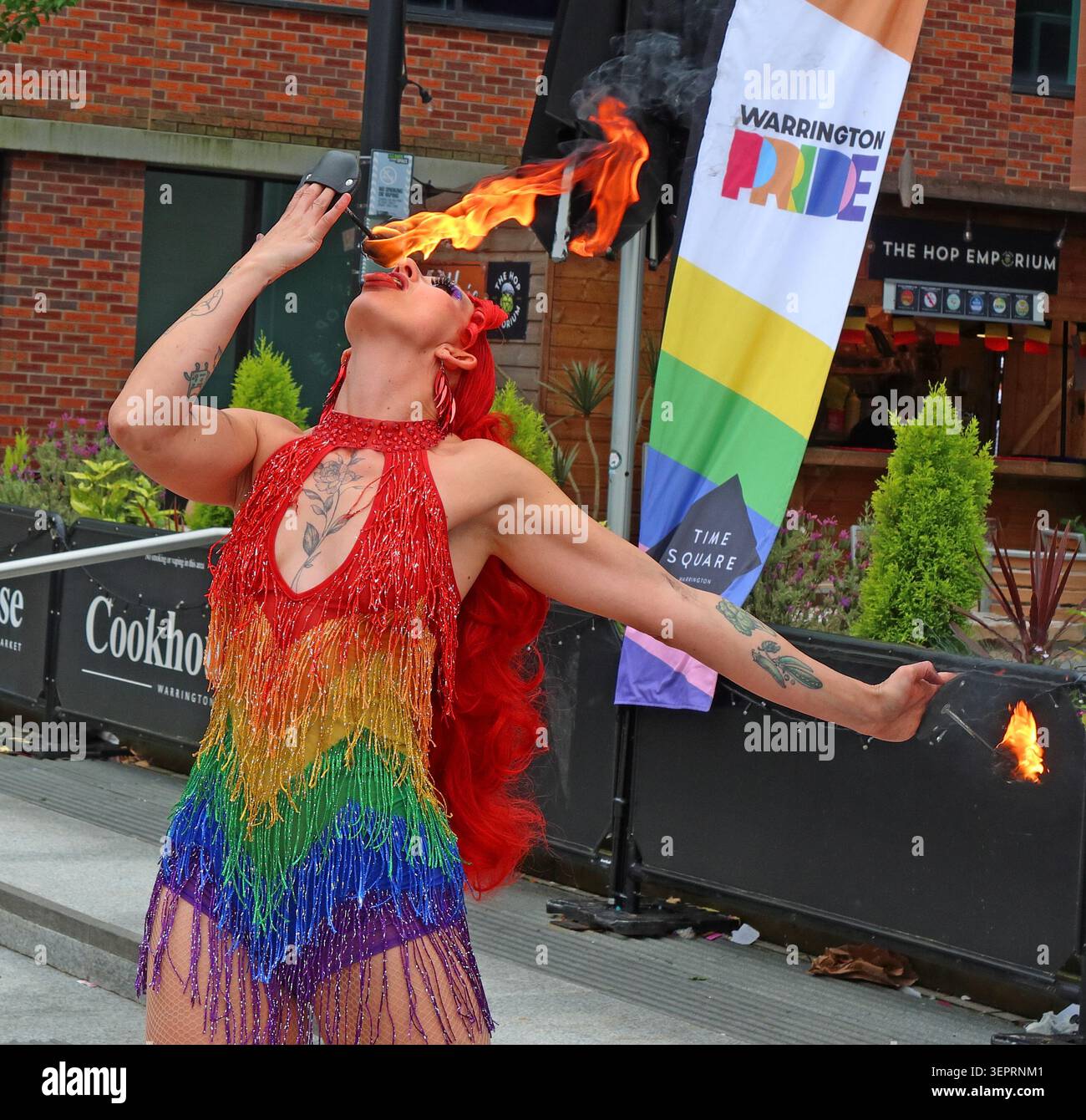 Tattooed fire performer entertains at Warrington town centre Pride 2026 in rainbow costume, beside a LBGTQ+ branded Pride banner. Stock Photo