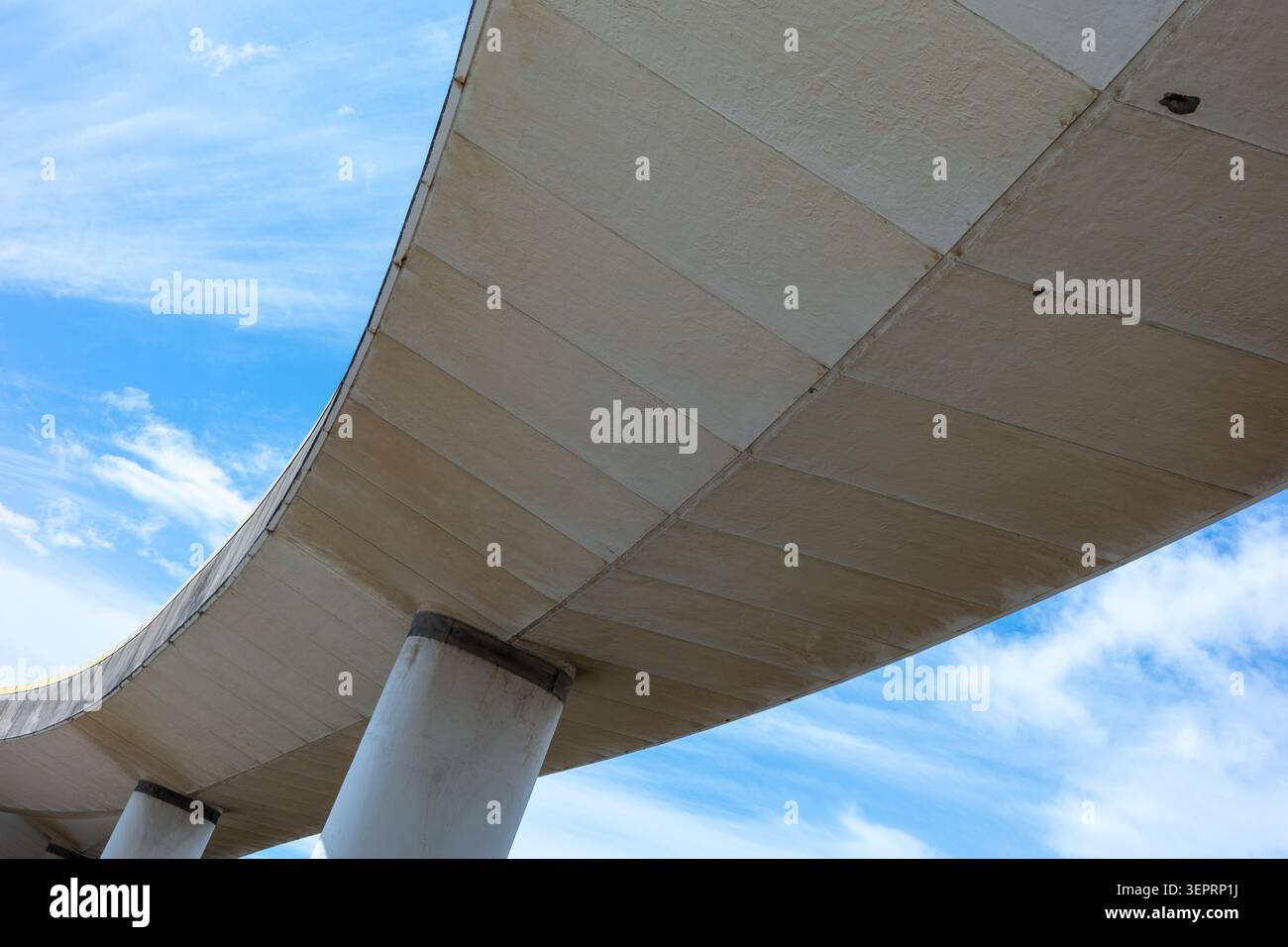 The scene shows a curved concrete structure viewed from below under a blue sky filled with soft white clouds. This structure is part of an urban lands Stock Photo