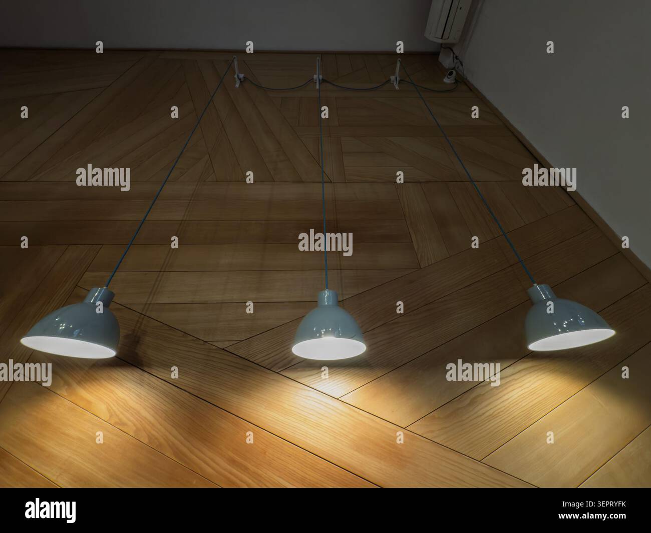 Three light blue pendant lamps hang low and illuminated over a light wooden floor with a geometric parquet pattern, showing indoor lighting, warm shad Stock Photo