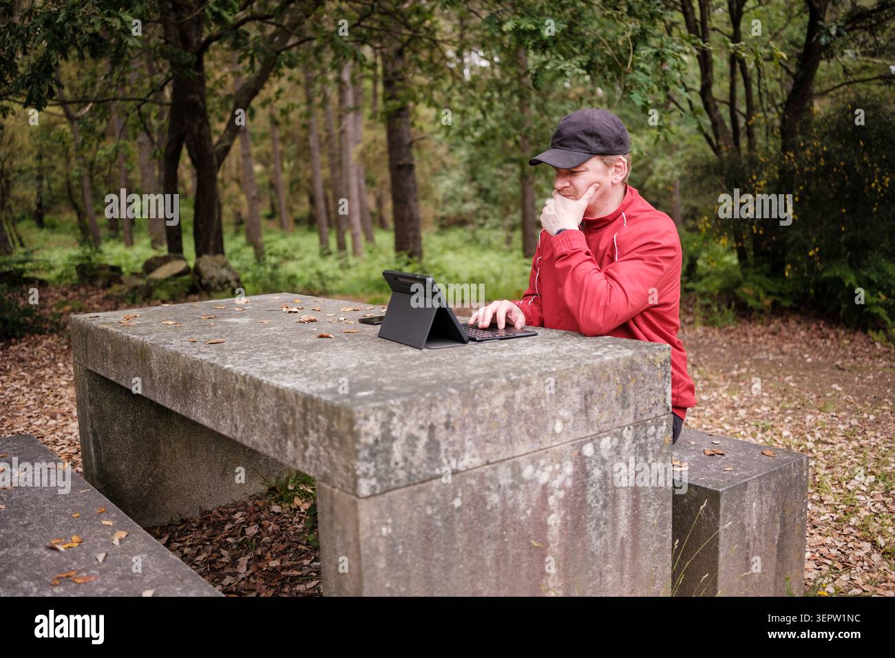 Man using a digital tablet with keyboard at a stone picnic table in a forest Stock Photo