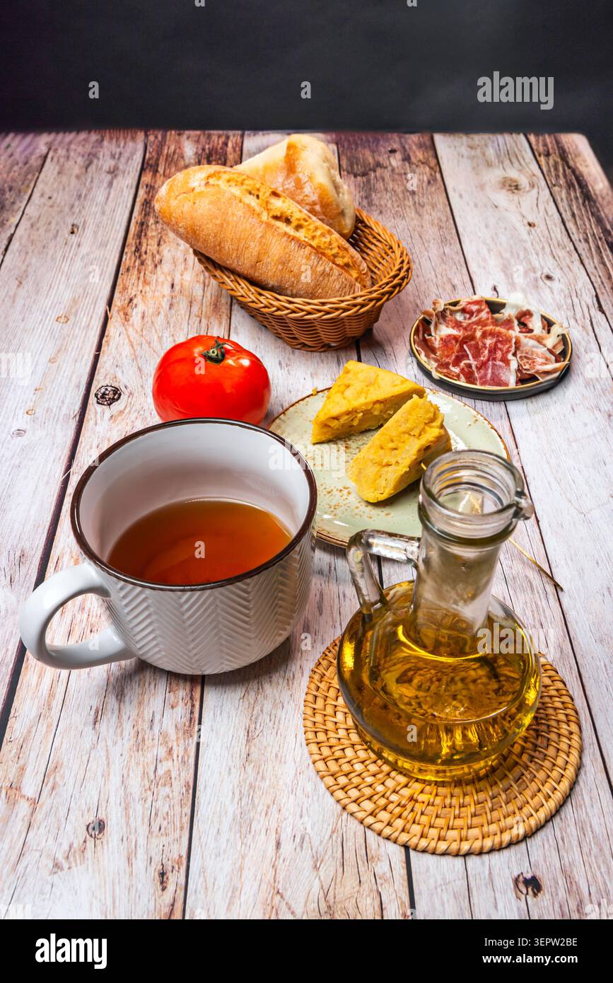 Glass olive oil cruet and white enamel pitcher beside bread basket, ham, tortilla chunks Stock Photo