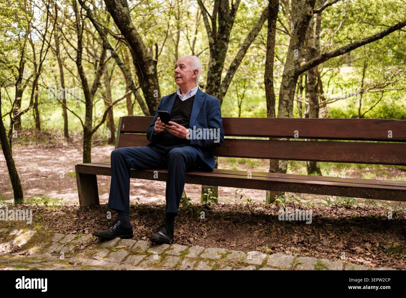 Senior man sitting on a park bench using a smartphone while looking away Stock Photo