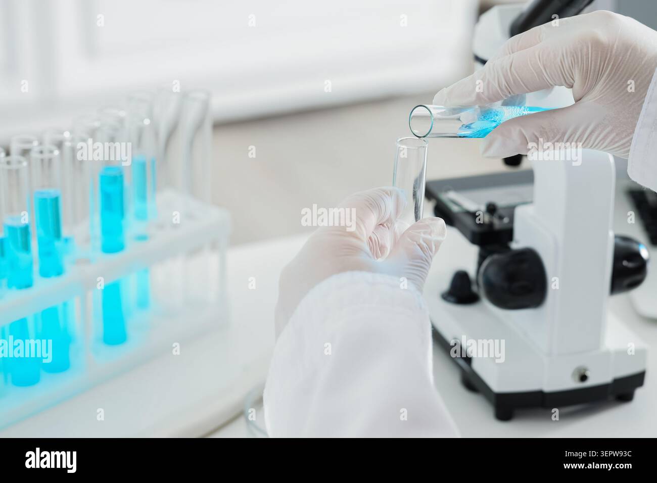 Scientist pouring blue liquid from test tube into another test tube in laboratory with microscope and rack Stock Photo