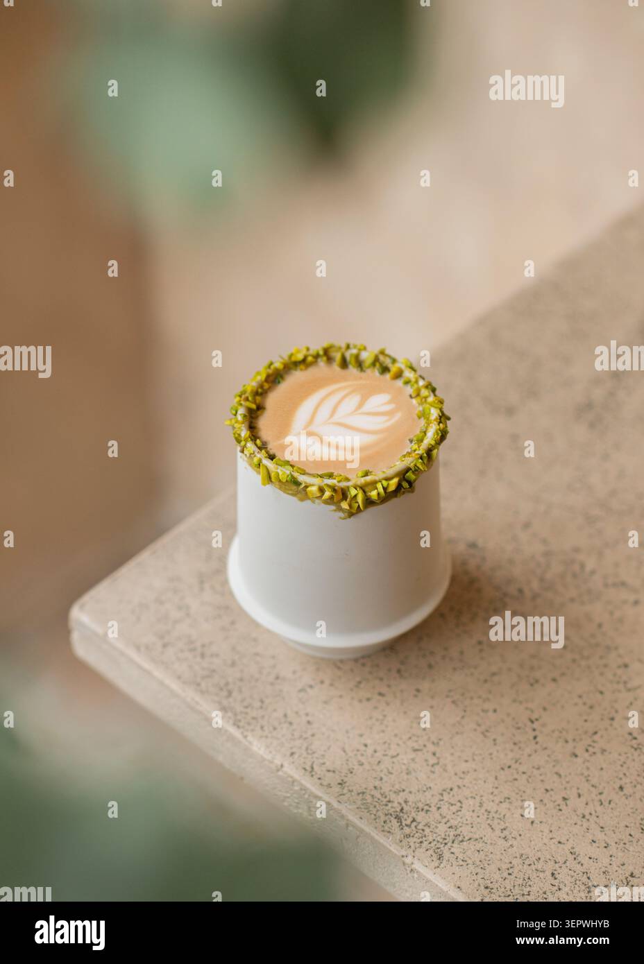 A hot pistachio latte showcasing detailed leaf art in a white ceramic cup with a crushed pistachio rim, set on a textured stone surface. Stock Photo