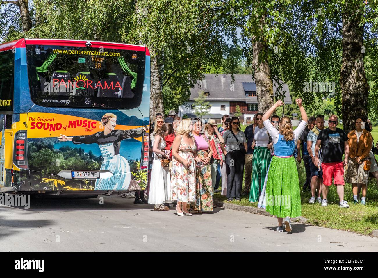 Tour guide Stephanie Hinterbuchner in a dirndl cheers on the tour group with her fist raised in front of the rear of a Panorama Tours coach on Fürstenweg, near Leopoldskron Castle in Salzburg; the coach bears the inscription ‘The Original Sound of Music Tour’. Salzburg Panorama Tours is the officially licensed operator of this city tour of the film locations from *The Sound of Music* (1965). Salzburg, Austria Stock Photo