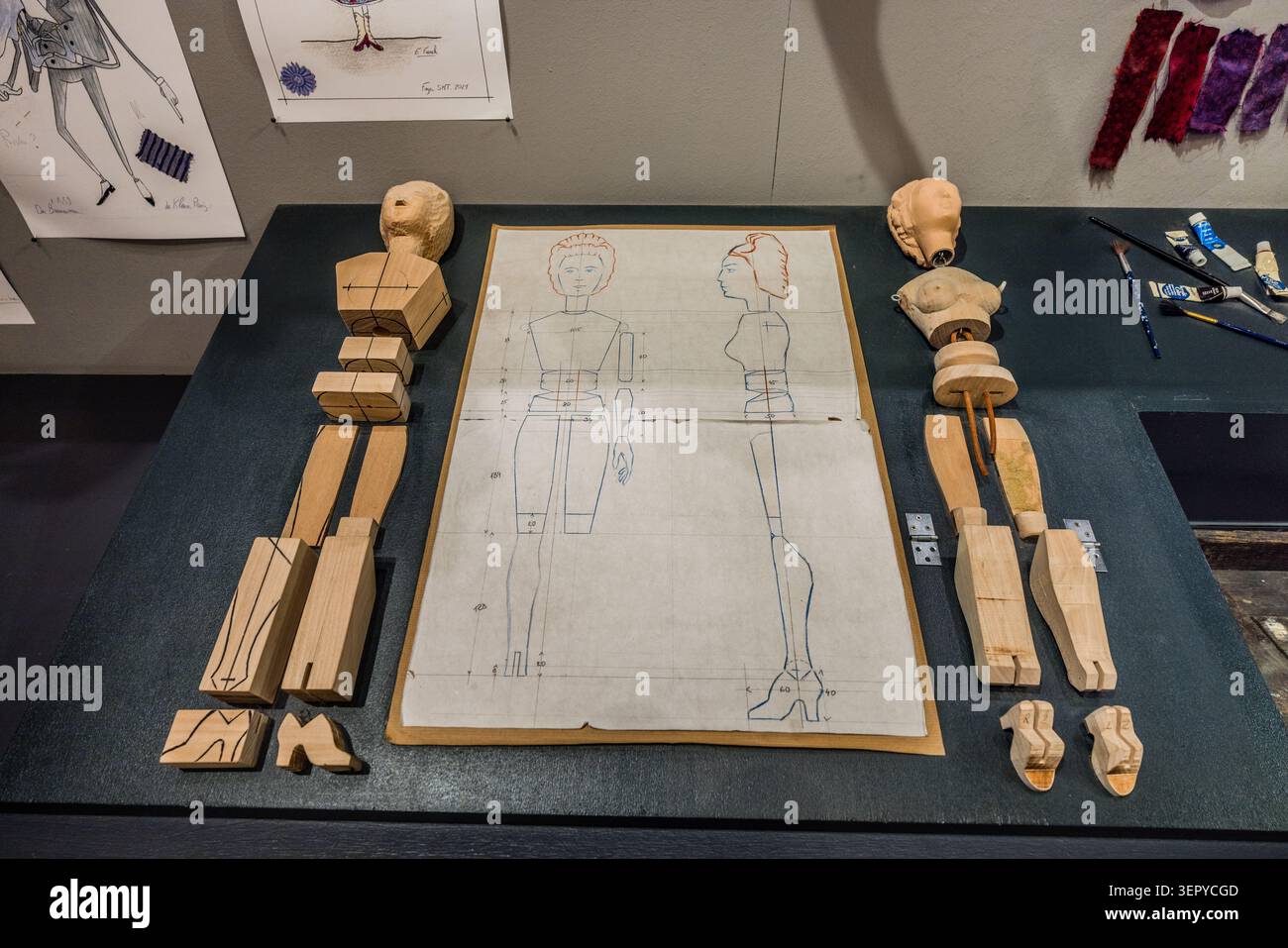 Workshop exhibition at the Salzburg Marionette Theatre: The dismantled wooden parts of a marionette – head, torso and limbs – lie on a workbench alongside a scale construction plan. Costume designs and colour samples can be seen in the background. The production of a marionette using the Salzburg method requires specialised craftsmanship. Salzburg, Austria Stock Photo
