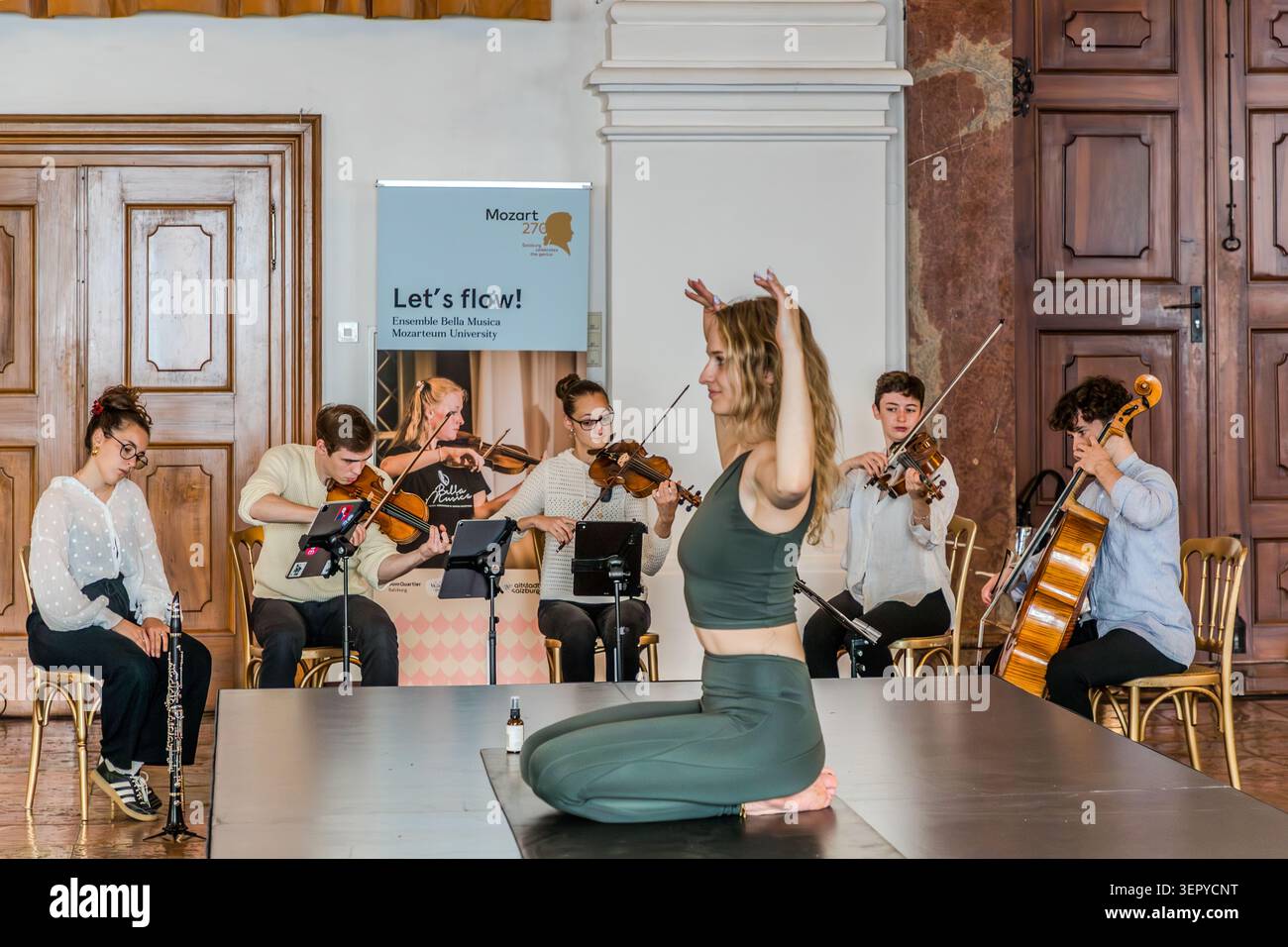 A yoga teacher in a balancing pose in front of the Bella Musica ensemble from the Mozarteum University in Salzburg – clarinet, several violins and cello; in the background, the banner ‘Mozart 270 / Let's flow!’. DomQuartier, Salzburg, Austria Stock Photo