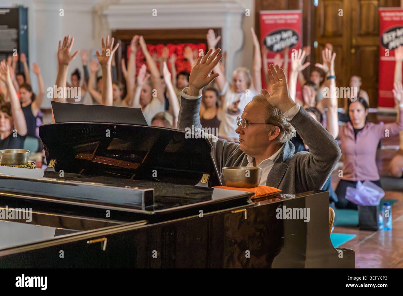 Pianist Stefan David Hummel at the concert grand piano raises his arms; behind him, dozens of female yoga participants also raise their hands in the air; singing bowls are placed on the piano. The event combined piano accompaniment, sound meditation and yoga as part of the Mozart 270 programme. DomQuartier, Salzburg, Austria Stock Photo