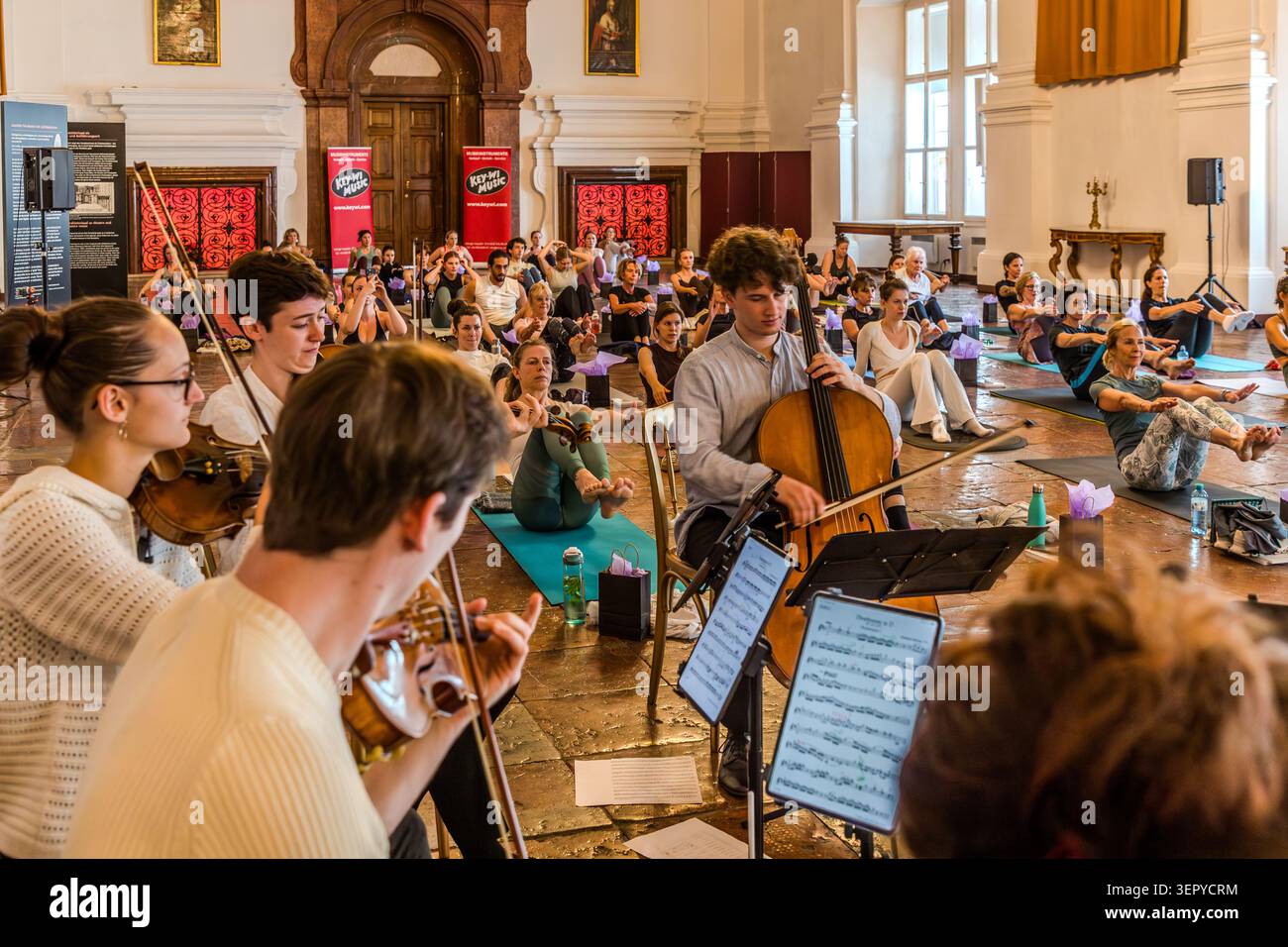 Members of the Ensemble Bella Musica from the Mozarteum University in Salzburg play string instruments in the foreground, whilst yoga participants sit on mats in the background; red exhibition banners adorn the wall. DomQuartier, Salzburg, Austria Stock Photo