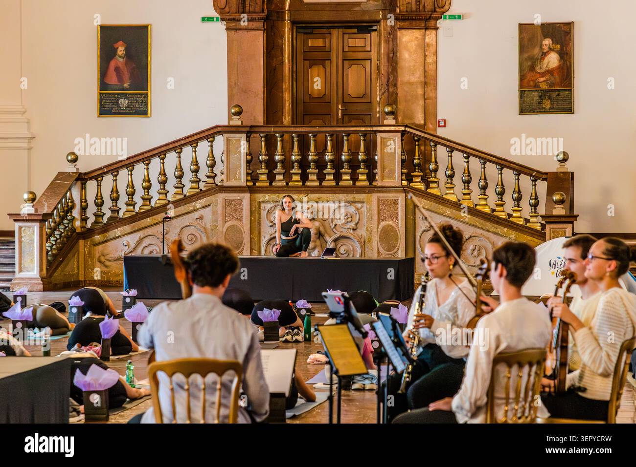 A panoramic view of the Baroque ballroom at the Residenz: yoga participants in the child’s pose on mats; on the right, the Bella Musica ensemble with a clarinet and violin; in the background, the grand Baroque staircase; a white SalzburgerLand promotional tent stands at the edge. DomQuartier, Salzburg, Austria Stock Photo