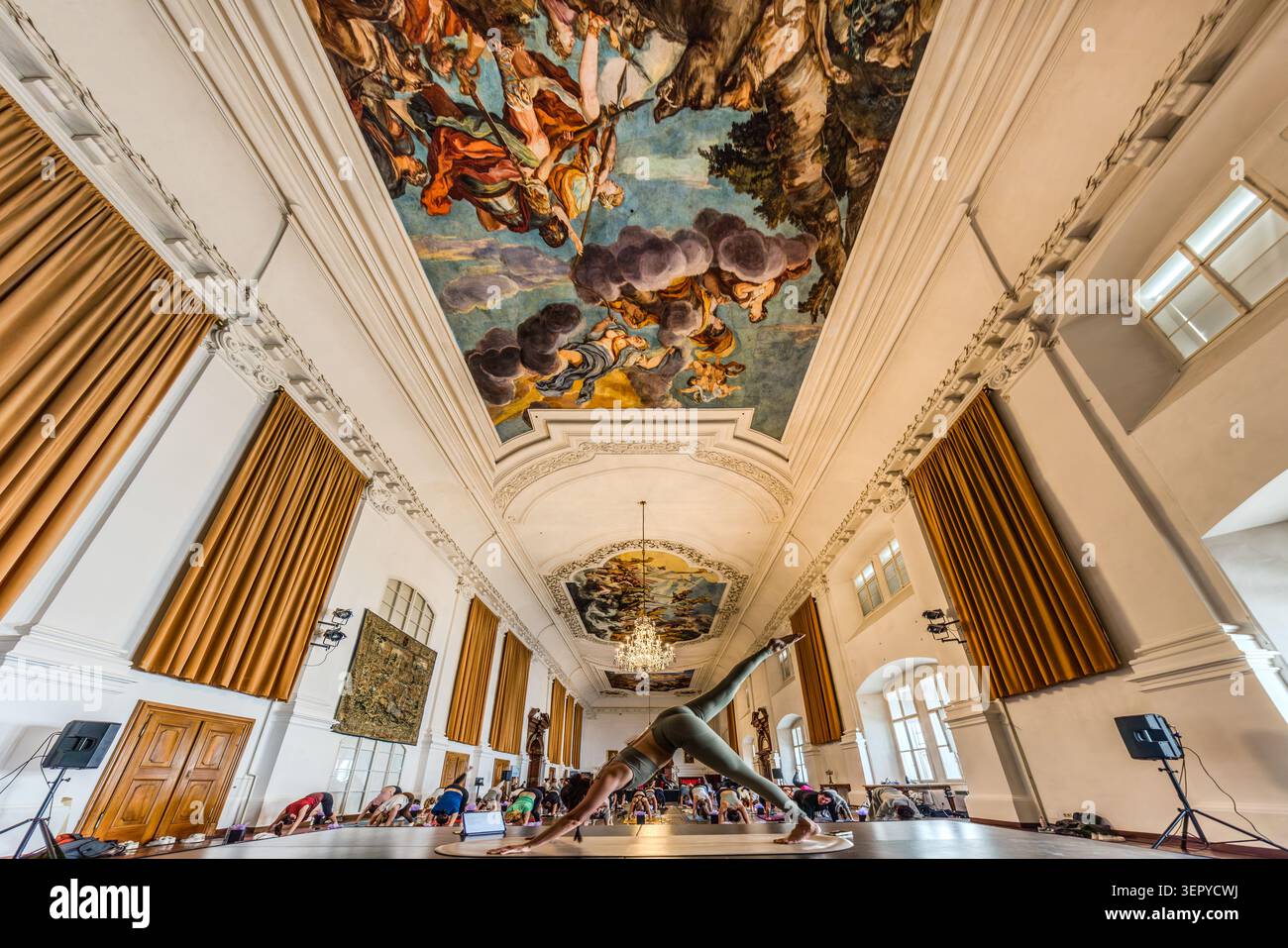 The Baroque ceiling fresco in the ballroom of the Archiepiscopal Residence (DomQuartier Salzburg) dominates the scene; in the foreground, a yoga instructor demonstrates a standing pose, whilst behind her the participants practise on mats. The hall is one of the stately reception rooms of the former residence of the Archbishops of Salzburg. Salzburg, Austria Stock Photo