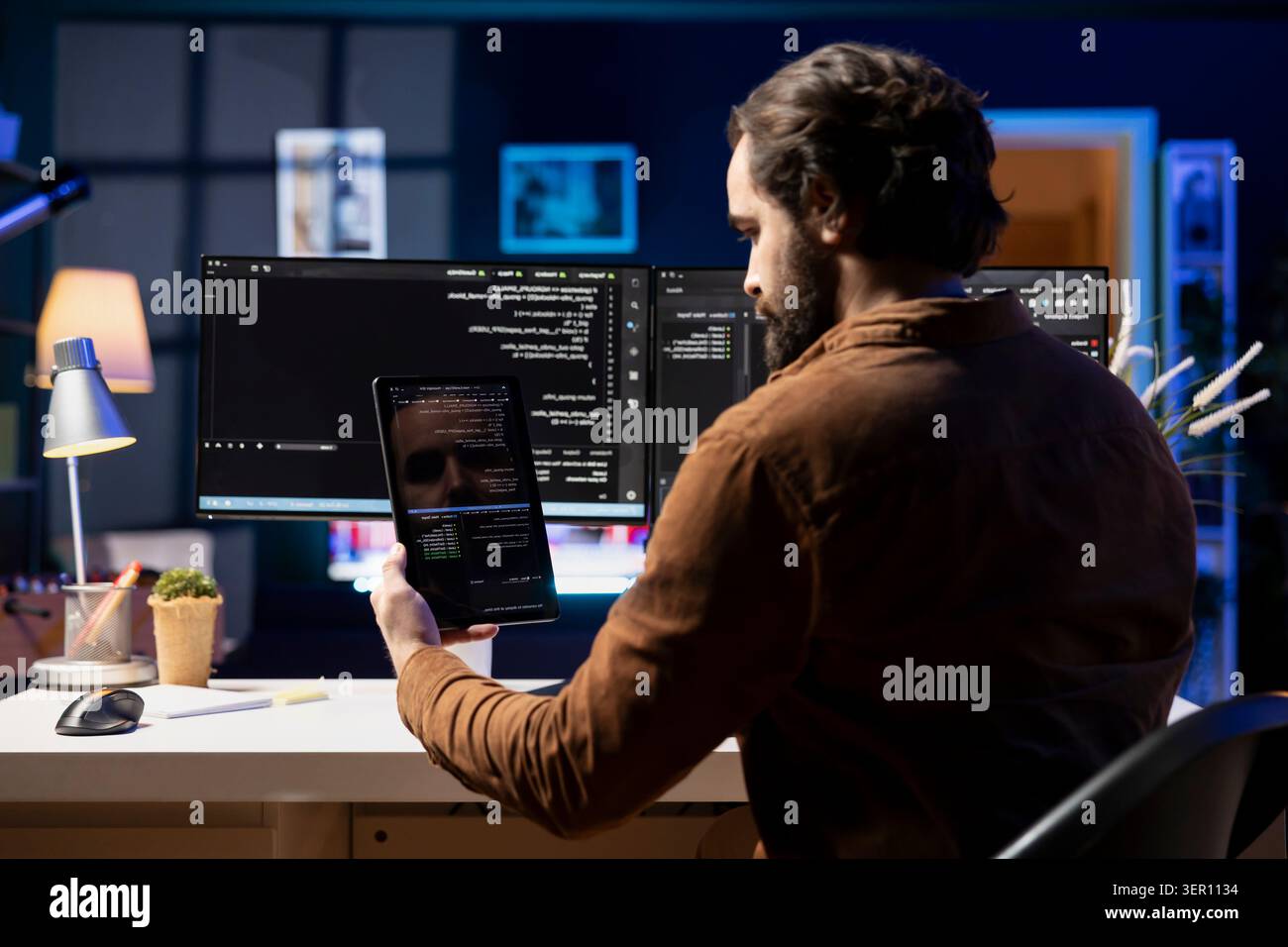 Programmer doing software debugging using tablet, checking glitches in lines of code displayed on computer monitors. Man working from home fixing coding issues using portable digital device. Stock Photo