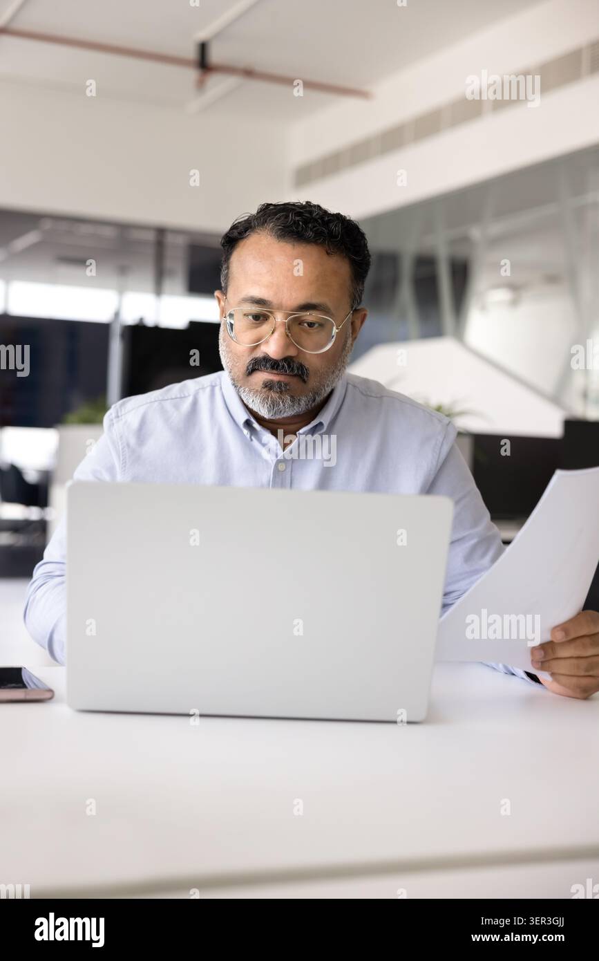 Focused Indian man comparing printed documents with digital data Stock Photo