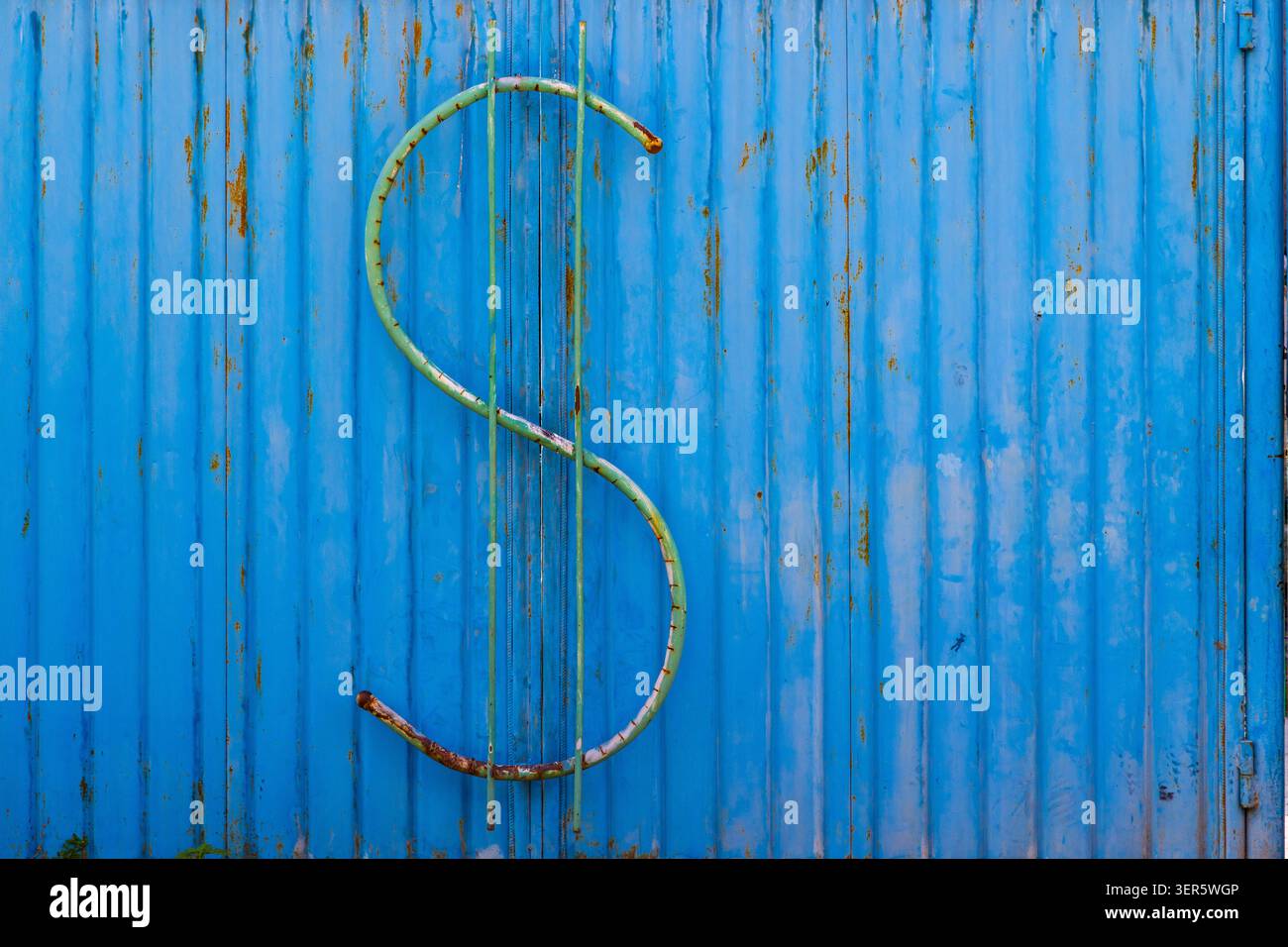 Large green dollar sign sculpture mounted on bright blue corrugated metal. Rust and wear on sculpture highlight age against textured industrial backgr Stock Photo