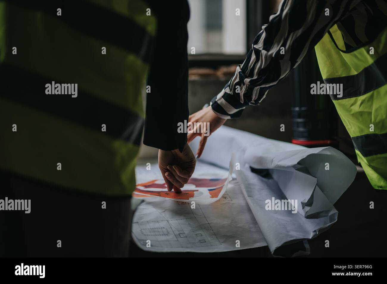 Construction professionals reviewing blueprints on site while wearing high-visibility safety vests Stock Photo
