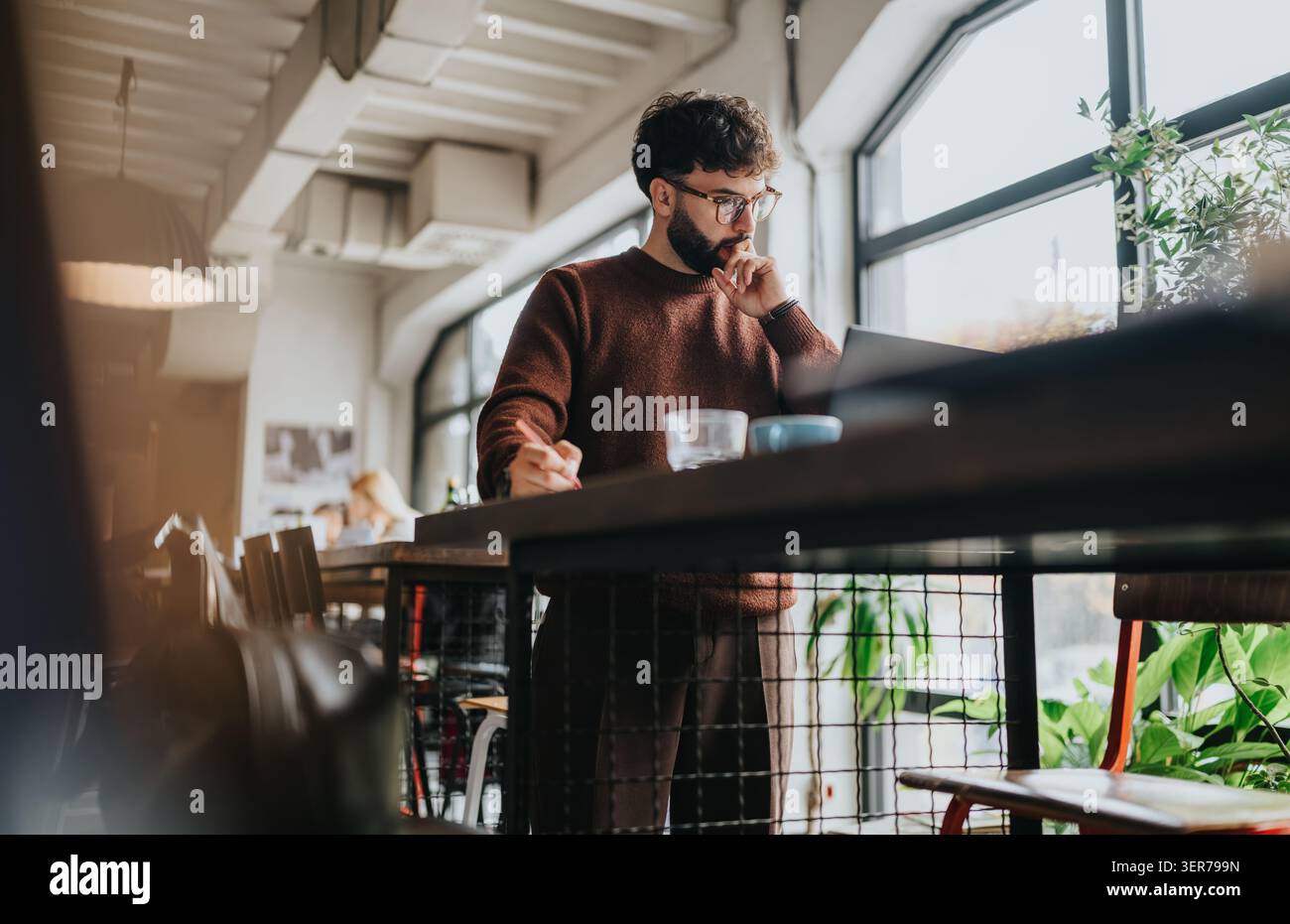 Young businessman working on laptop and taking notes while standing in a modern cafe workspace Stock Photo