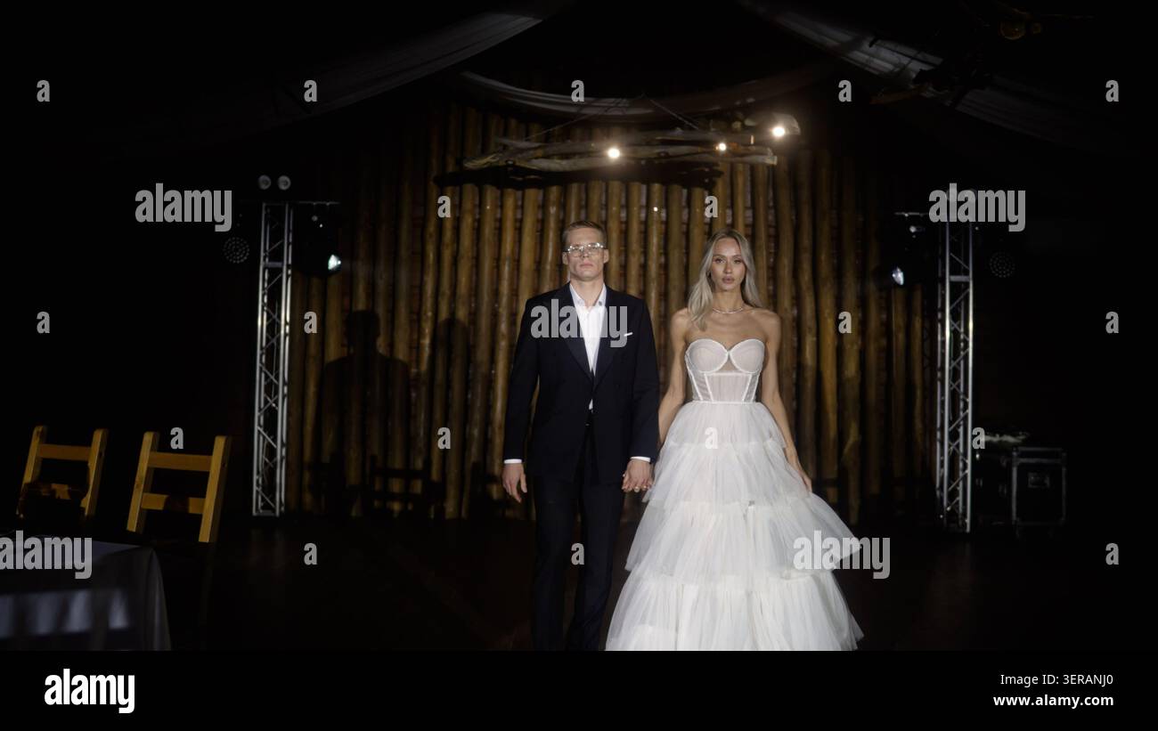Elegant young couple, man in black tuxedo, woman in white tiered gown, walk hand in hand. Dramatic lighting highlights their confident pose against da Stock Photo