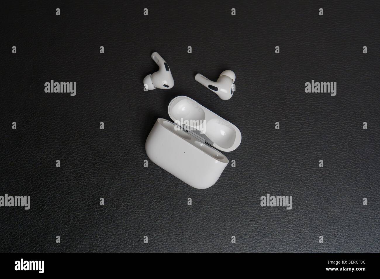 White earbuds rest beside open charging case. Surface has fine, dark textured grain. Device shows clean, minimalist design. Earpieces sit open, ready Stock Photo
