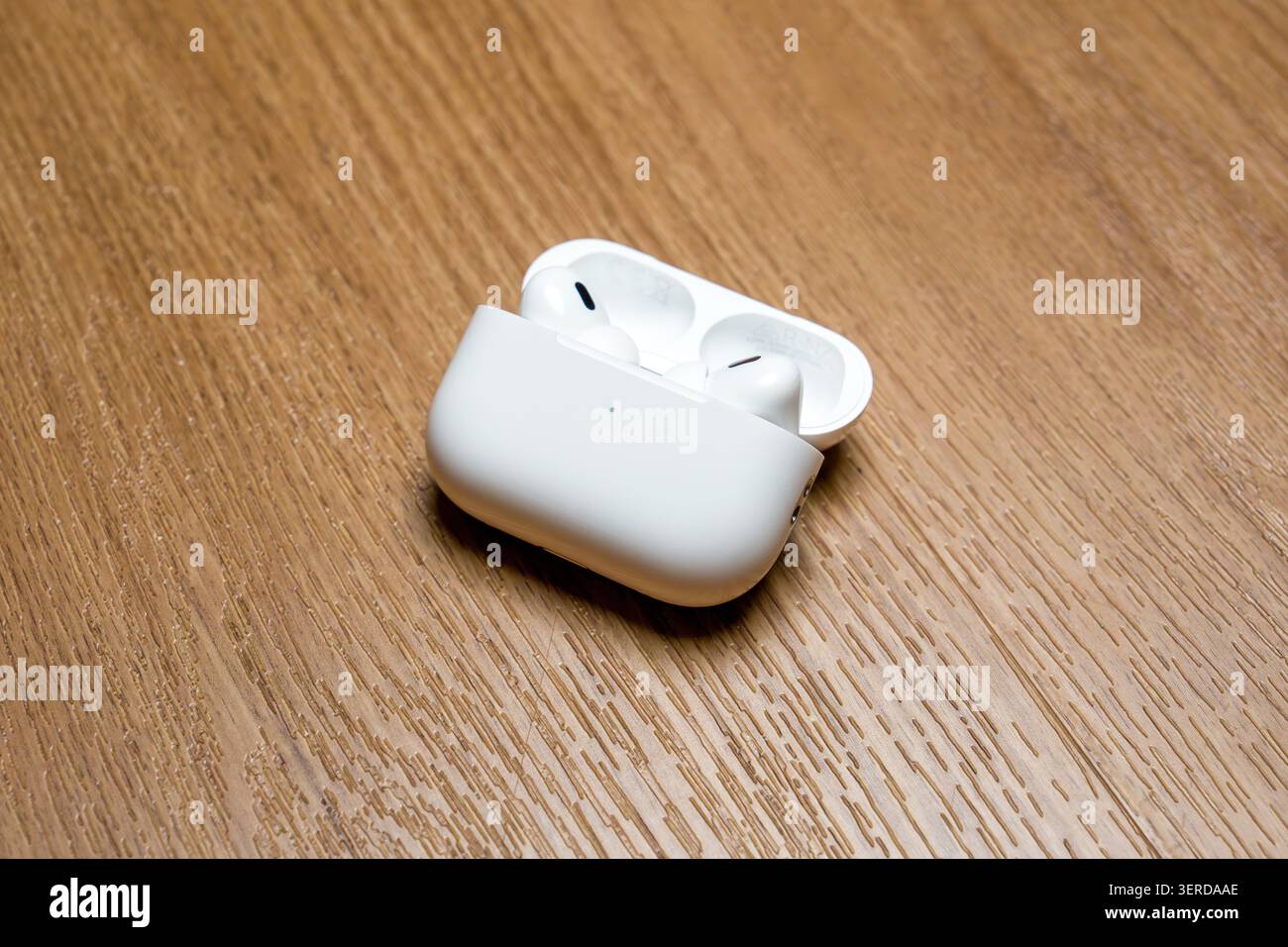 White earbuds rest in open charging case. Case sits on light wood surface. Minimalist, clean design visible. Soft lighting highlights product details. Stock Photo