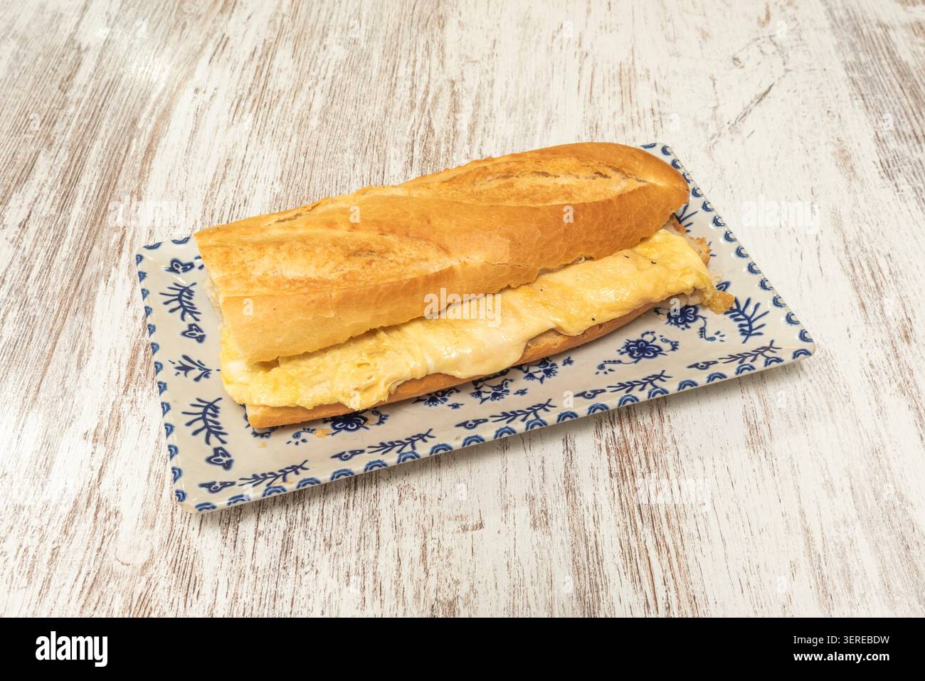 A crusty baguette sandwich filled with thick folded French omelette sits on a blue and white patterned rectangular plate over wood. Stock Photo