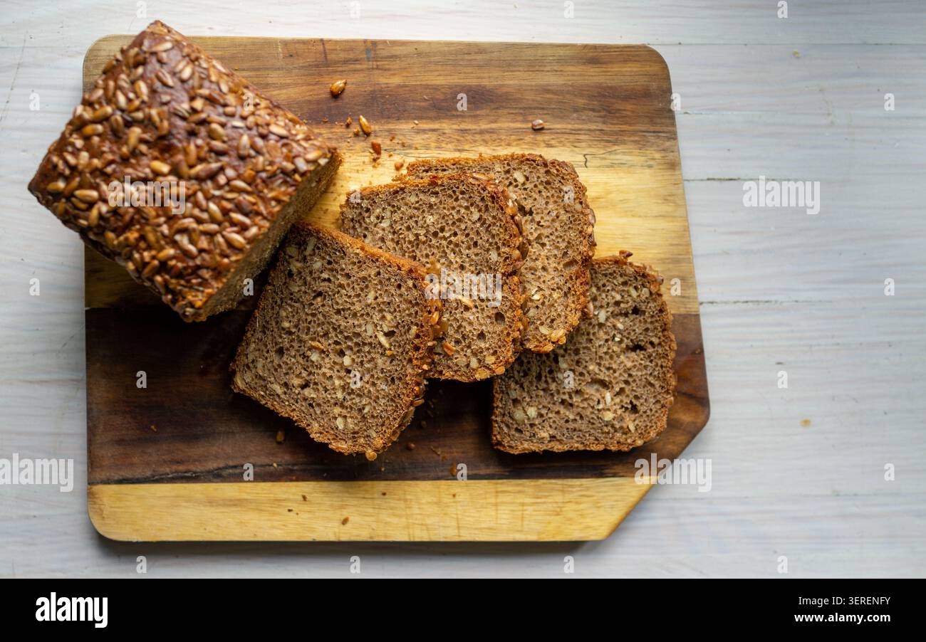 Top-down view of sliced whole grain loaf topped with sunflower seeds arranged on rustic wooden cutting board placed on pale tabletop visible crumb tex Stock Photo