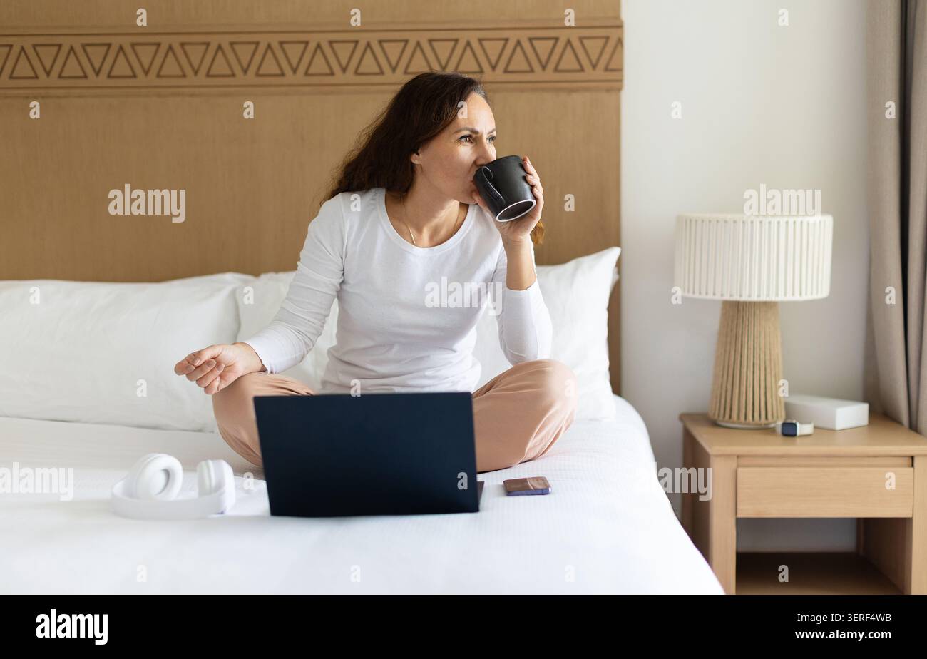Woman drinking coffee while using laptop on hotel bed, working remotely Stock Photo