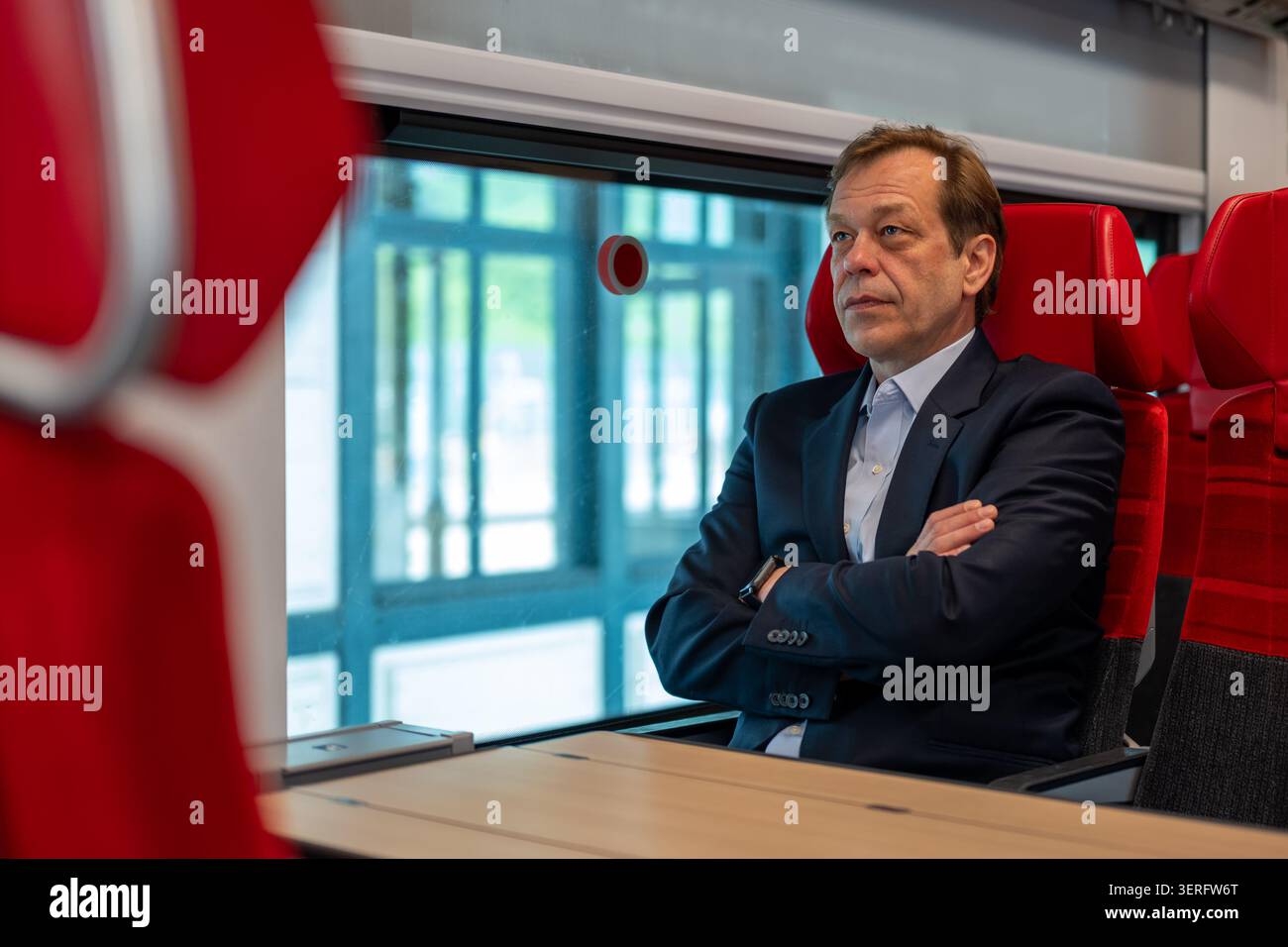 Middle aged man sits in a train carriage with folded arms Stock Photo