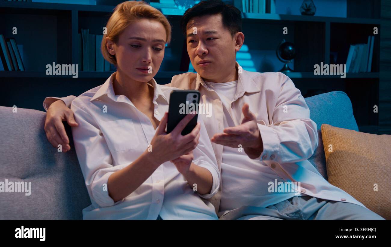 Multiethnic couple talking shopping using mobile phone choosing order has different opinions at evening at home. Asian man disagree with Caucasian Stock Photo