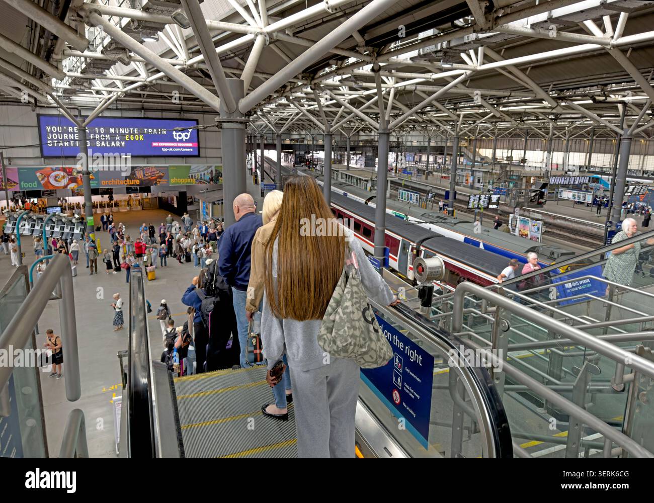 Passengers descend an escalator inside Leeds railway station, with platforms, trains and the busy concourse visible below. Stock Photo