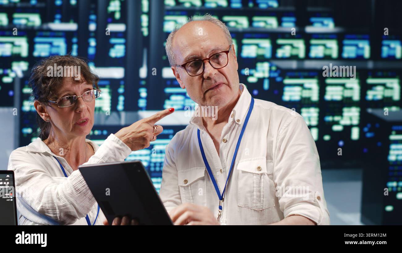 Software developers in high tech establishment, completing tasks on tablet while running scripts on laptop terminal, proactive programmers amidst server rows. Stock Photo
