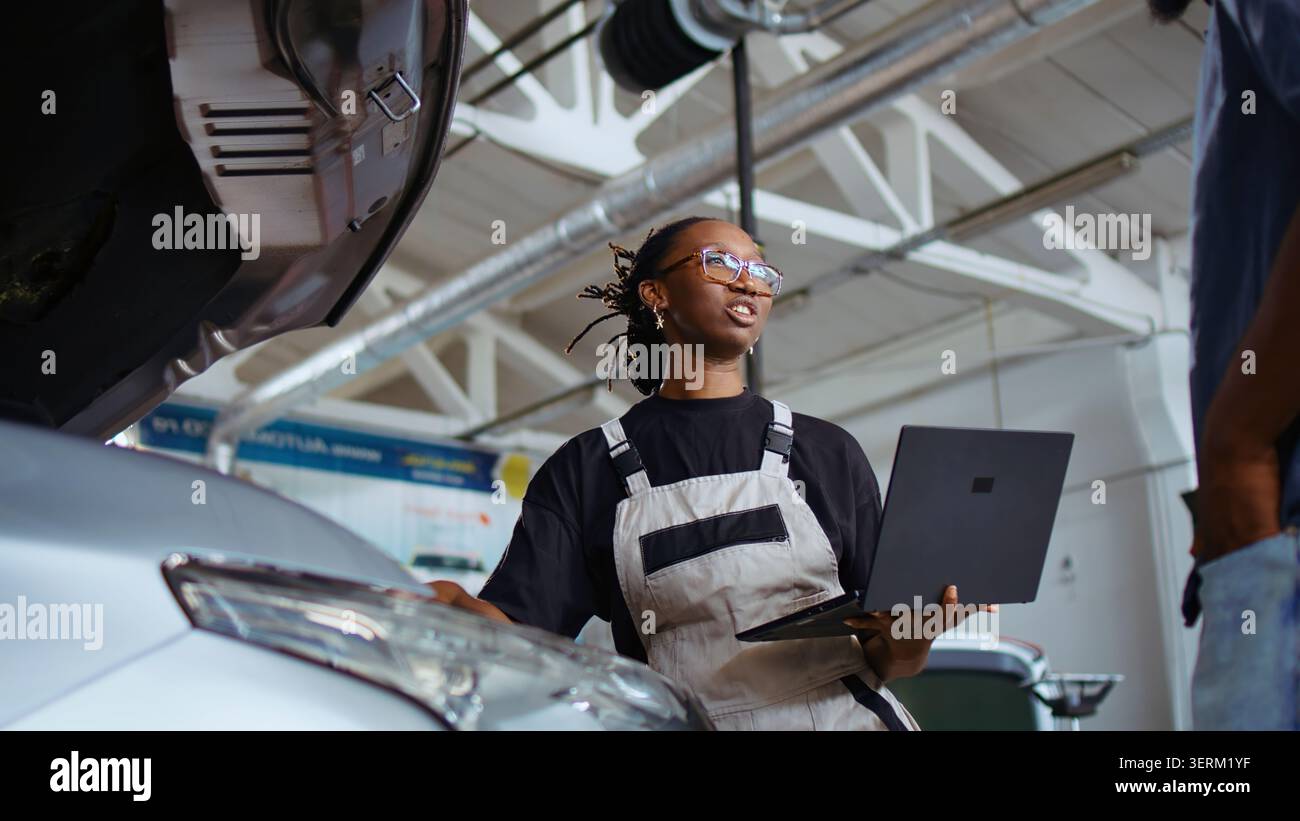 Experienced technician listens to customer in repair shop, using laptop to write down car modifications needed, professional in garage with client, listening to her requests, zoom out shot. Stock Photo