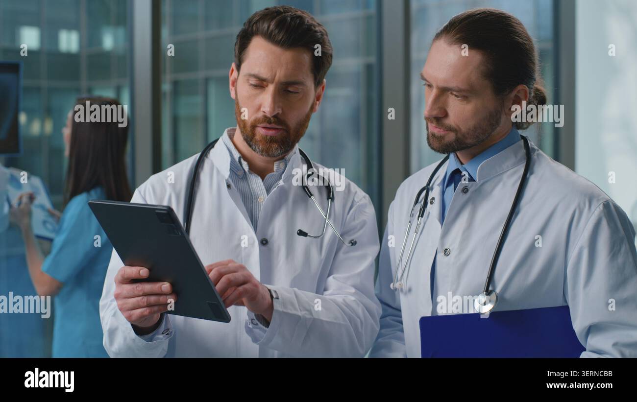 Two Caucasian doctors men talking analyzing health checkup results with digital tablet online medicine service in clinic. Coworkers physicians Stock Photo