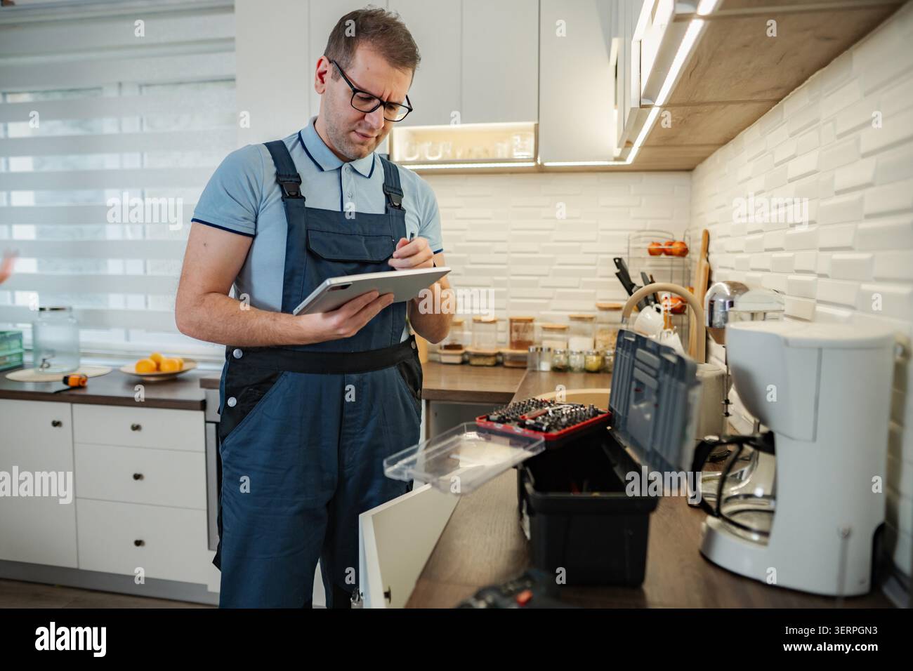 Handyman in overalls writing on a digital tablet while checking a kitchen Stock Photo