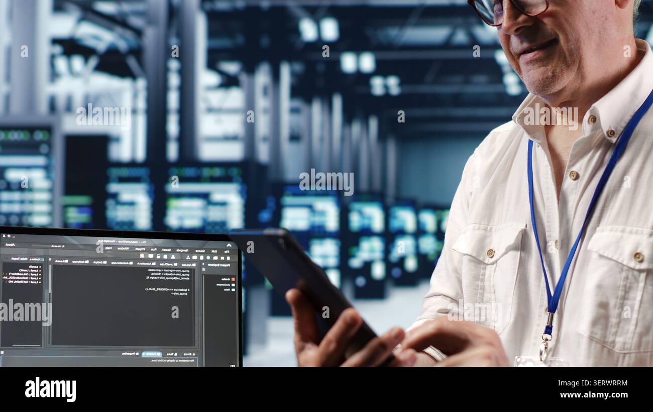 Senior technician in data center, solving tasks on tablet while running scripts on laptop terminal, with skilled technician amidst servers, accessing databases and manipulating lines of code. Stock Photo
