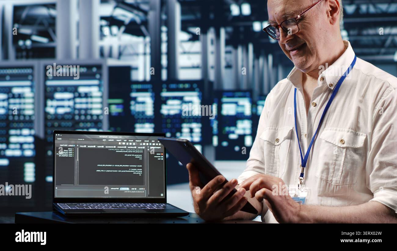 Elderly worker in data center, solving tasks on tablet while running scripts on laptop terminal, certified engineer amidst servers, accessing databases and manipulating lines of code. Stock Photo