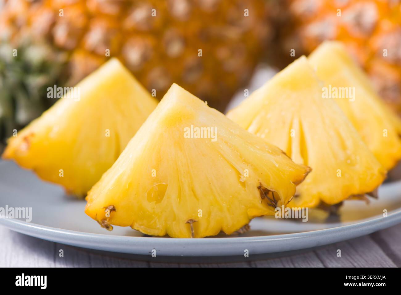 Pineapple slices are arranged on a white plate with whole and half pineapples in the background Stock Photo
