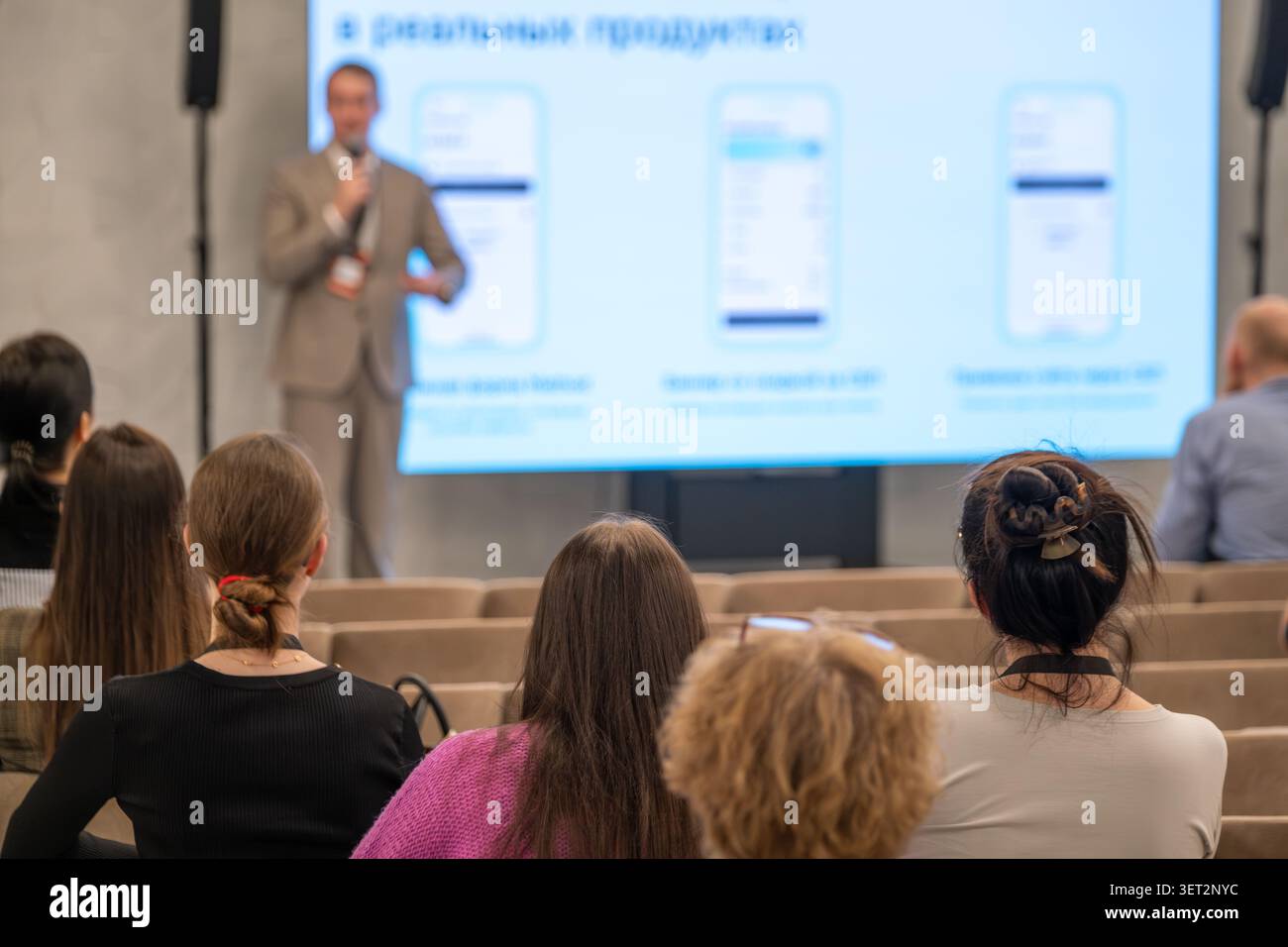 Business speaker presents slides to seated audience in modern conference room, attendees listen and take notes, professional seminar atmosphere. Stock Photo
