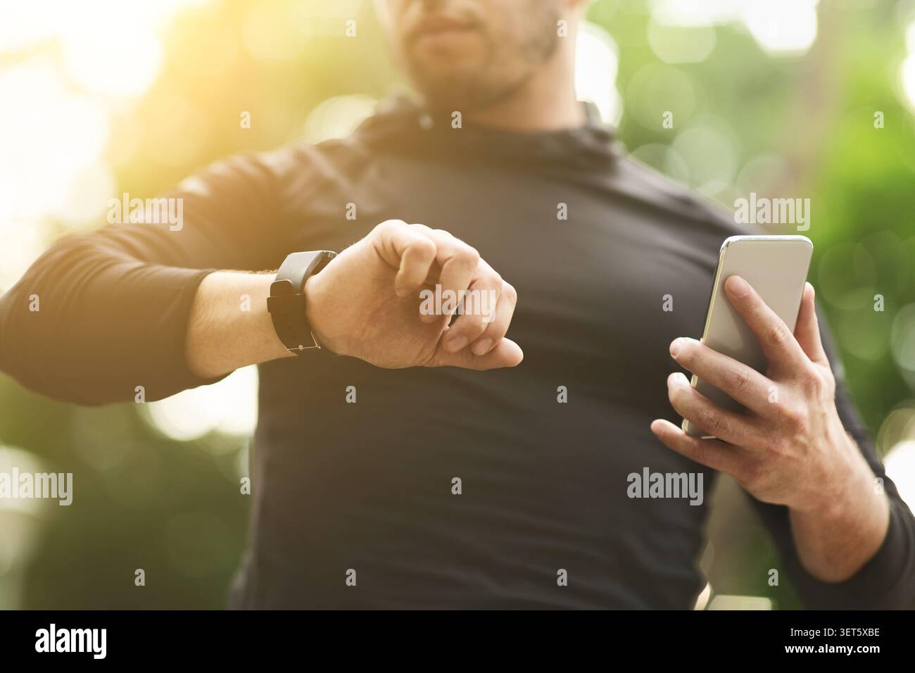 Man Checks Watch While Using Smartphone Outdoors in Sunlight Stock Photo