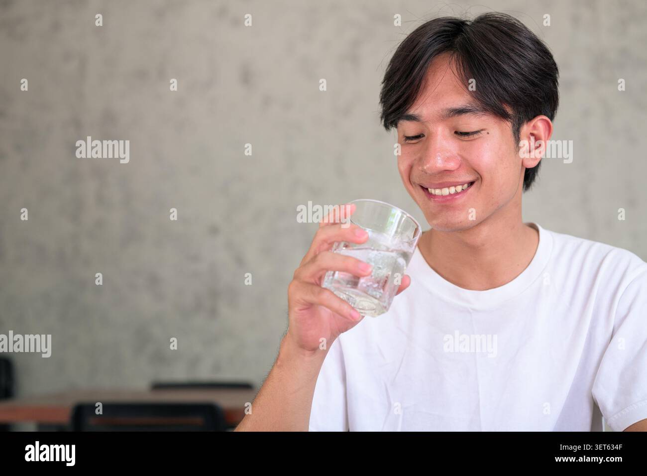Horizontal lifestyle photograph of a handsome young Southeast Asian Thai Isan non-binary man smiling happily while bringing a glass of water to his mo Stock Photo