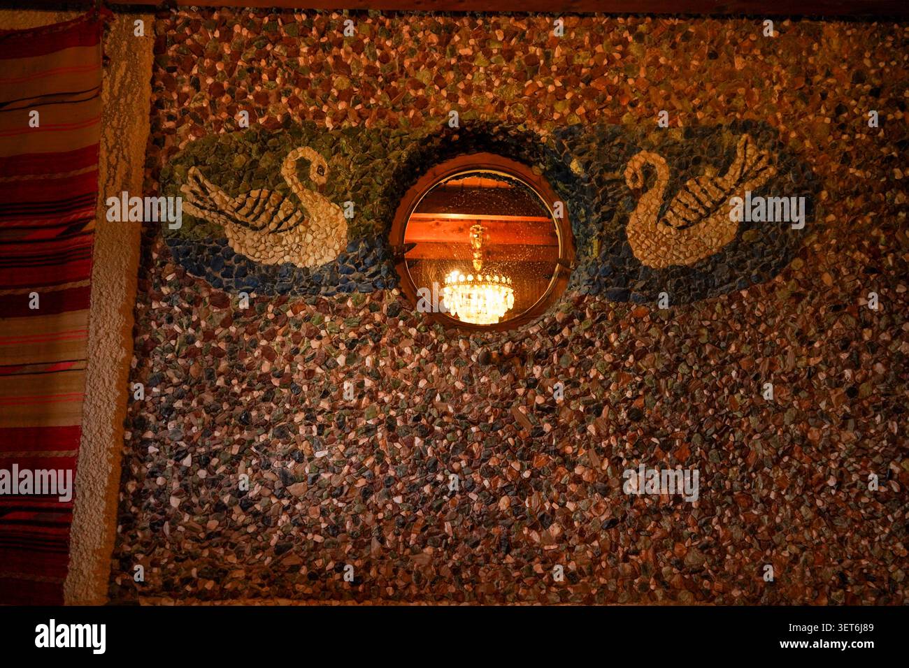 A unique textured mosaic pebble wall featuring two beautiful swans flanking a round window that reflects a glowing crystal chandelier inside. Stock Photo
