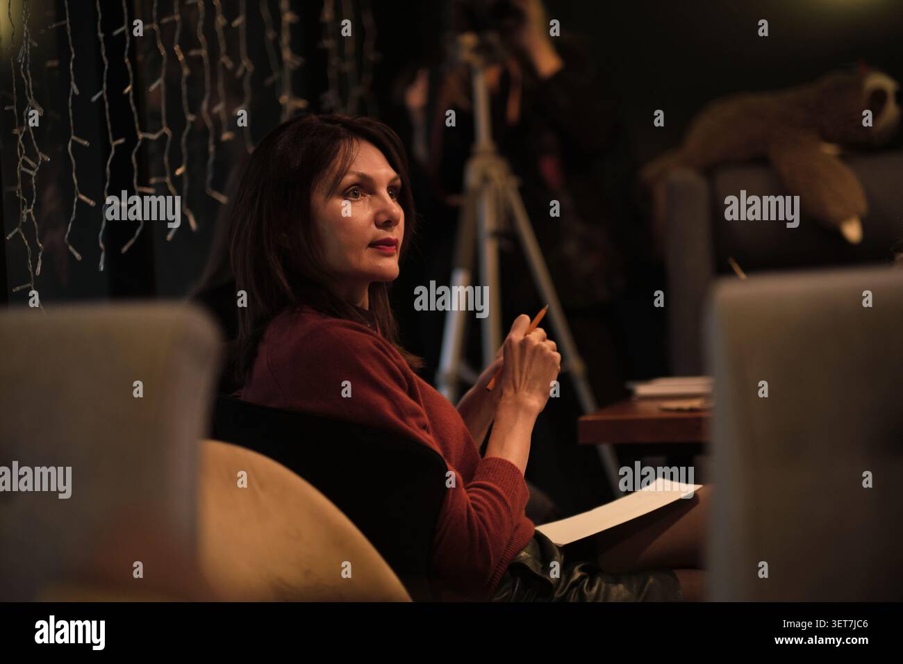Side view of a thoughtful woman in a red sweater sitting at a dim seminar, holding a pencil and notebook. Adult learning, workshops, and focus support Stock Photo