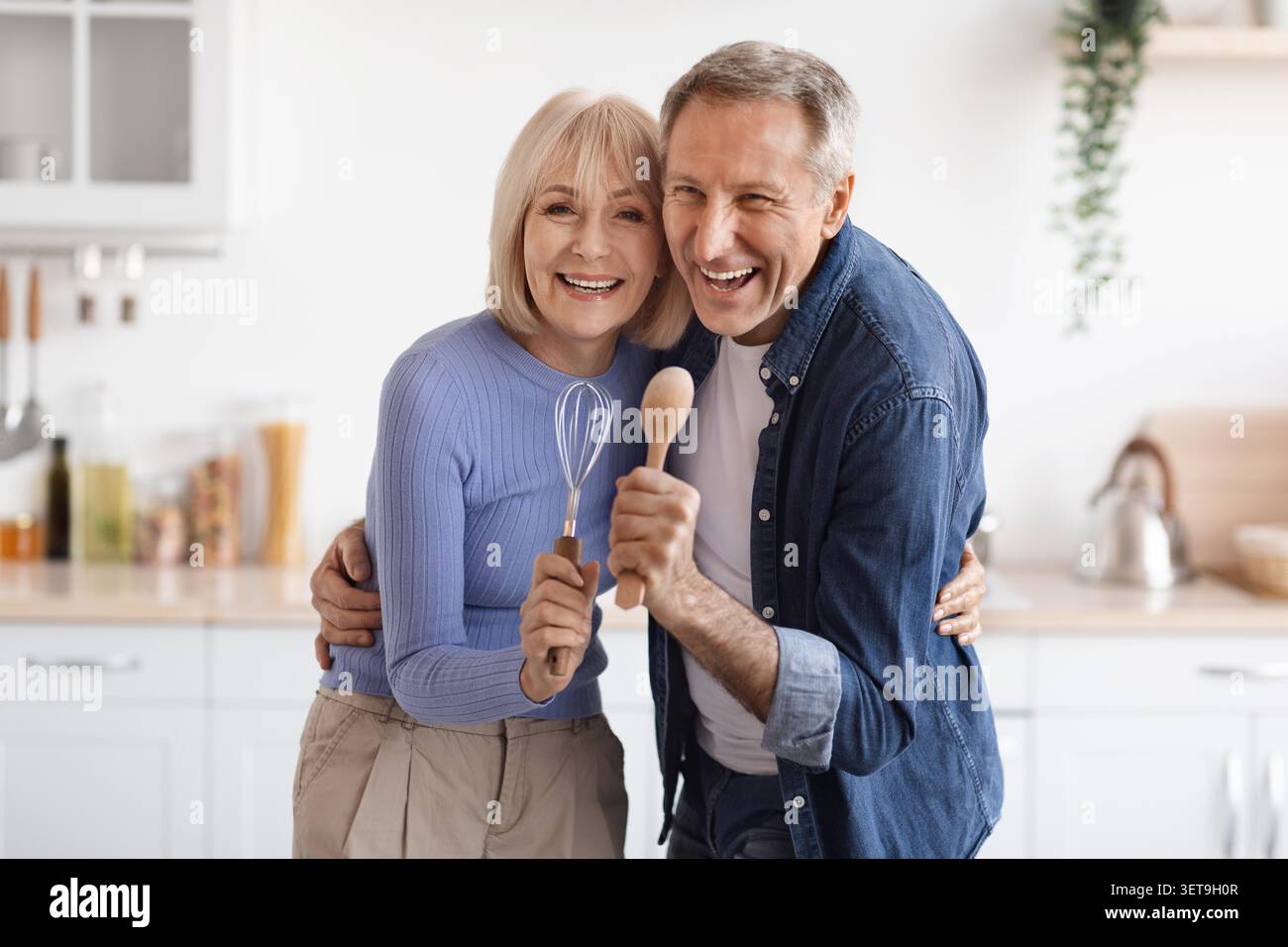 Happy Elderly Caucasian Couple Cooking Together In Kitchen, Smiling And Holding Utensils Stock Photo