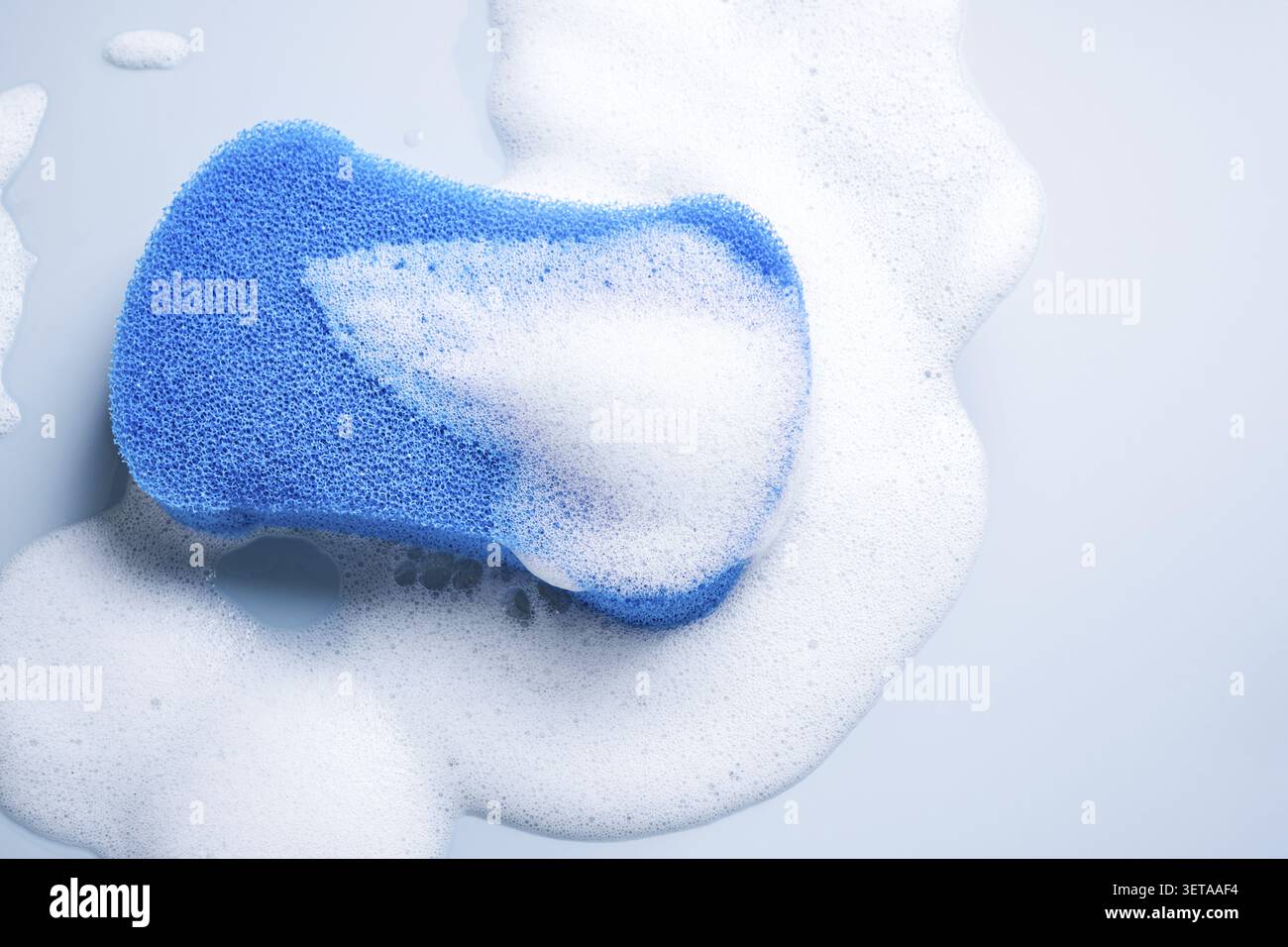Blue sponge surrounded by white soap foam on a light blue background Stock Photo