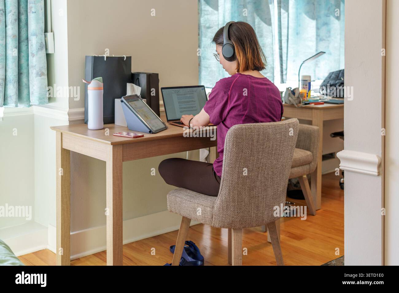 A dedicated student engages in online study using multiple devices in a comfortable workspace at home. Stock Photo