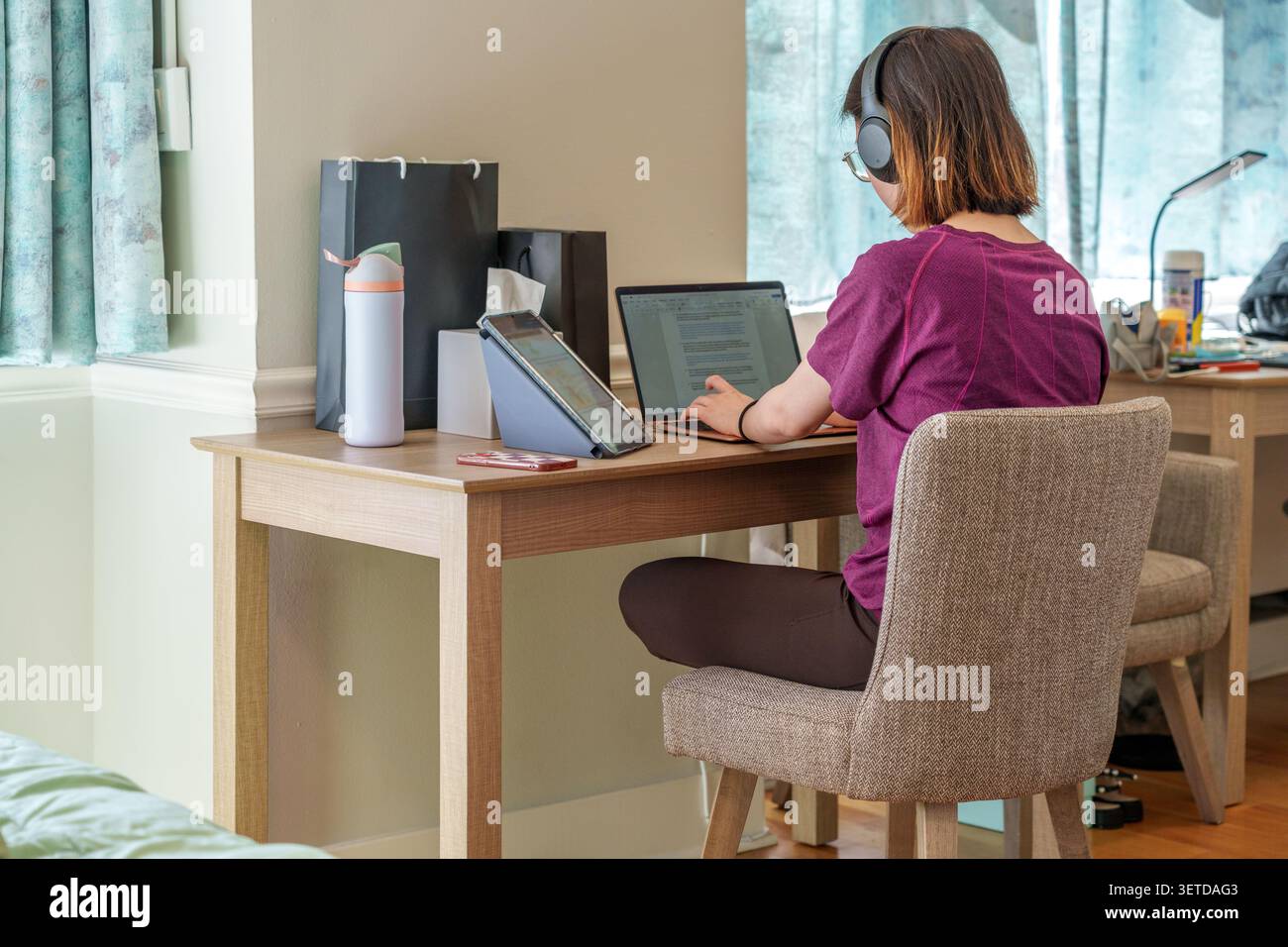 A dedicated student engages in online study using multiple devices in a comfortable workspace at home. Stock Photo