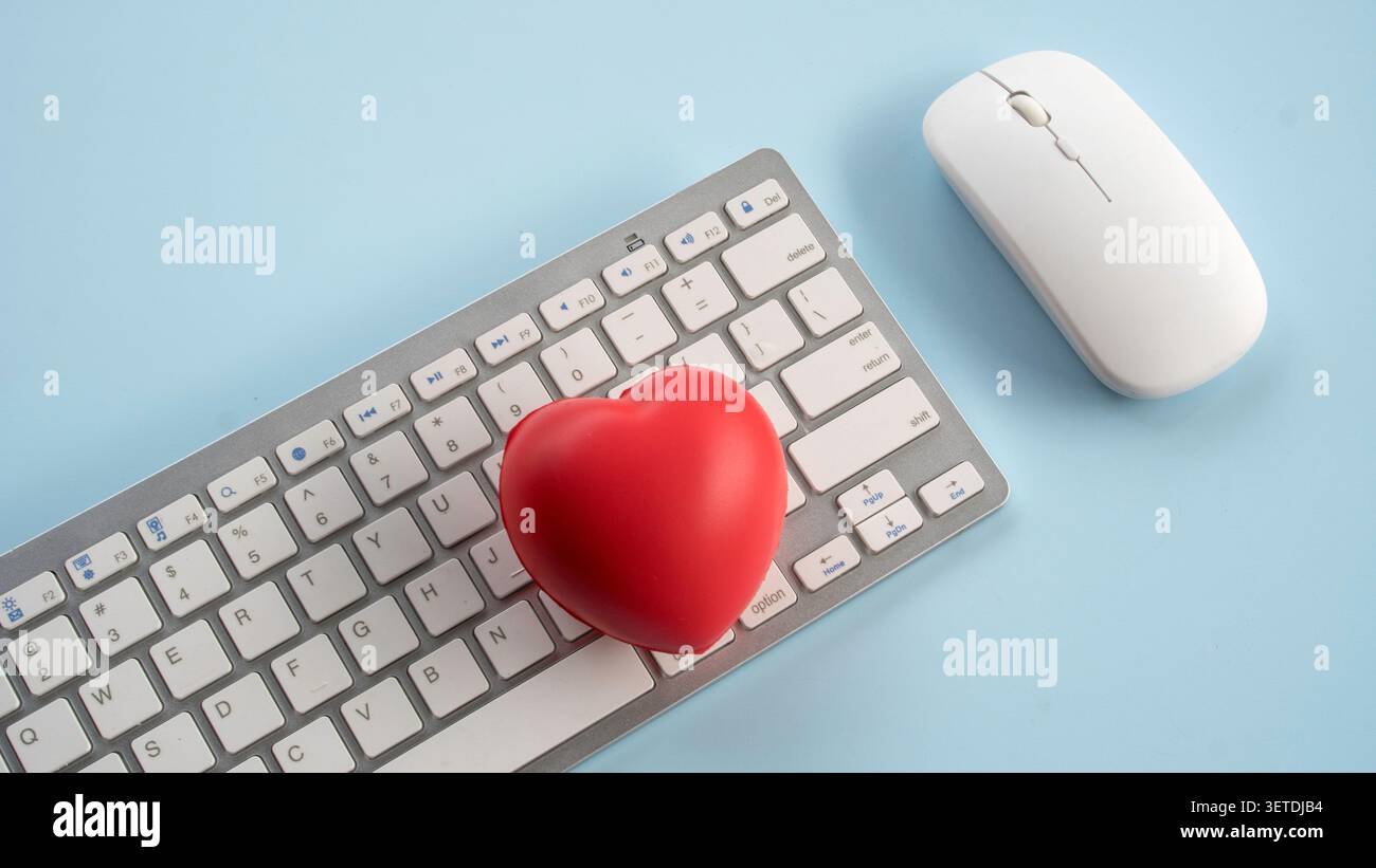 Red stress ball heart shape placed on computer keyboard next to wireless mouse on light blue desk representing healthcare technology online dating or Stock Photo