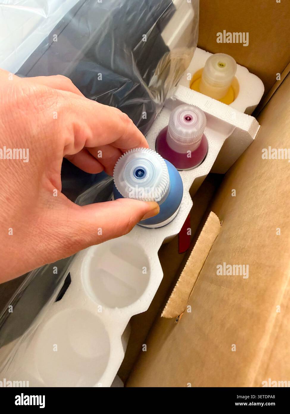 A person taking a blue ink bottle out of a cardboard shipping box containing a printer and a set of replacement ink refills. - Smartphone Captured Stock Image
