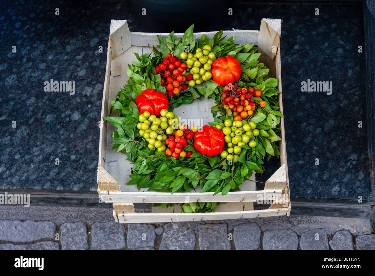 A decorative green leaf wreath is arranged inside a rustic wooden crate on a dark stone step. The arrangement is enriched with three small bumpy red p Stock Photo
