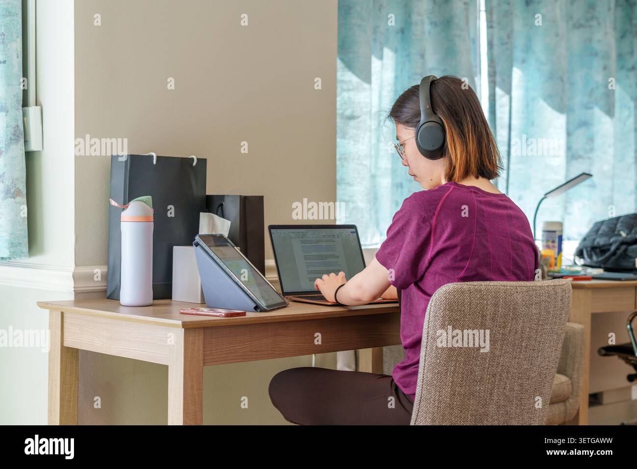 A dedicated student engages in online study using multiple devices in a comfortable workspace at home. Stock Photo