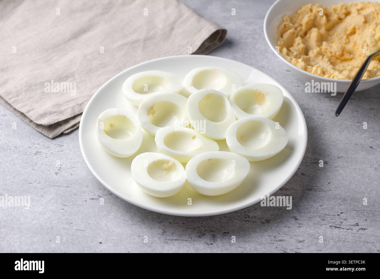 Hard boiled egg halves with yolks removed on a white plate, prepared for stuffed eggs recipe on a gray textured surface with soft natural light Stock Photo